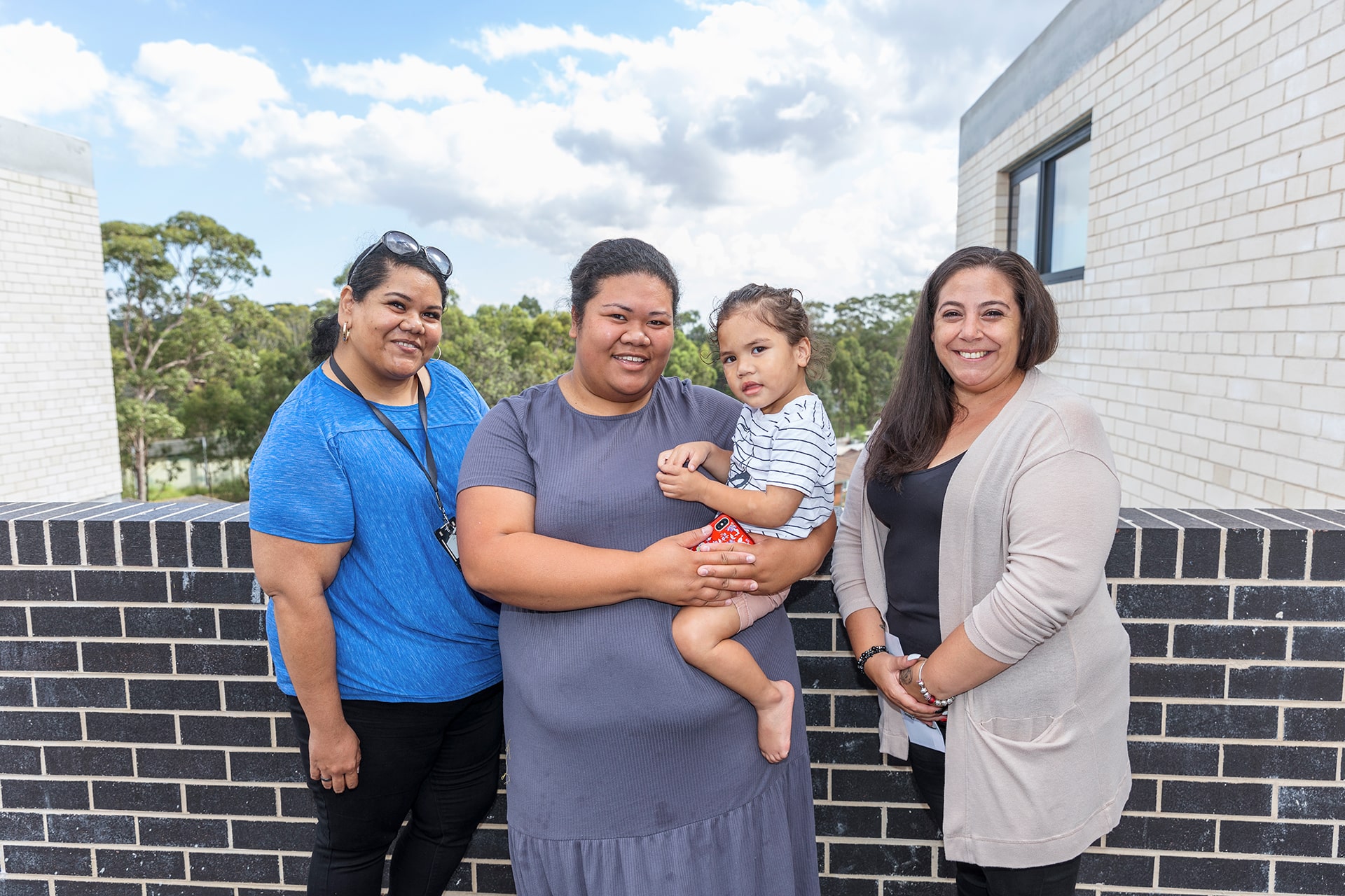 Three women on a balcony overlooking parkland, middle woman is holding a child