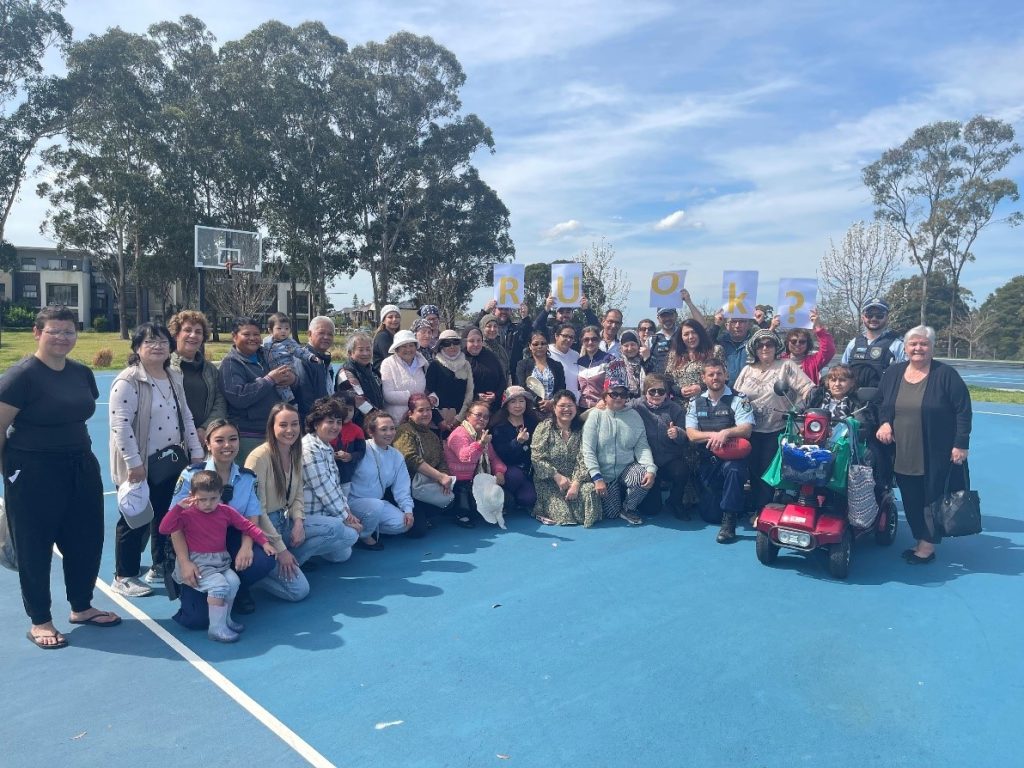 Dozens of people pose for a group photo on a sunny day in the park holding up a sign that says R U OK?