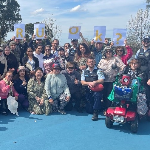 Dozens of people pose for a group photo on a sunny day in the park holding up a sign that says R U OK?
