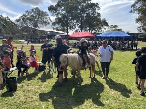 A child wearing a helmet rides a pony accompanied by adults. Other kids in helmets wait nearby