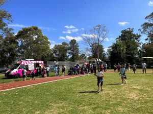 Children and adults queue in a sunny field at an ice cream truck