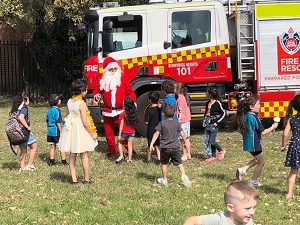 Santa stands in front of a fire truck parked in a sunny field with dozens of children coming to see him