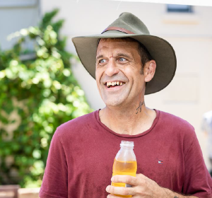 A man in a wide brim hat smiling outdoors and holding a bottle of apple juice