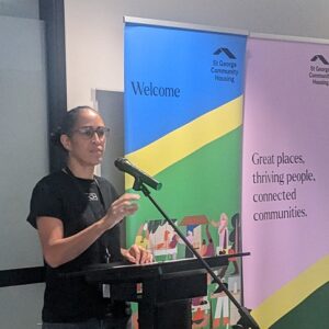 Stakeholder Engagement Manager Evelyn speaking from a lectern in front of pull up banners that say Welcome and Great Places, thriving people, connected communities.